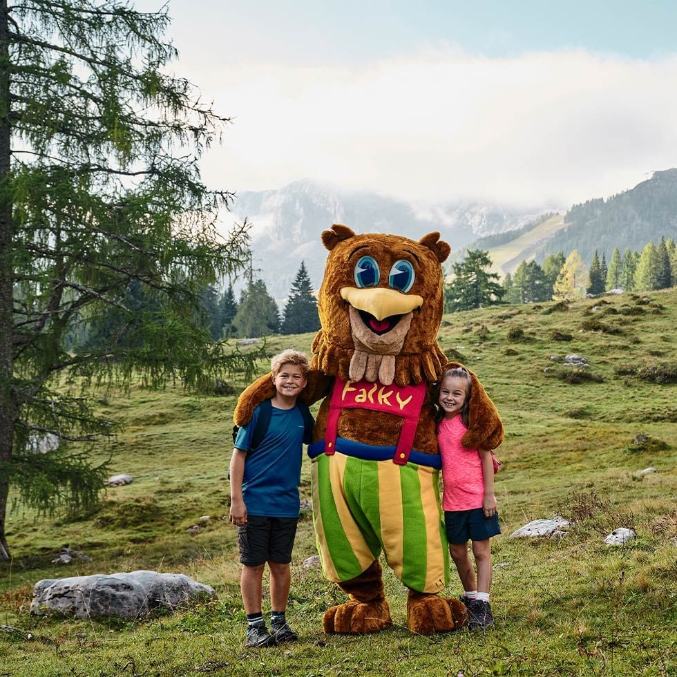 Zwei Kinder und ein Bär-Maskottchen stehen in einer Almlandschaft vor einem Bergpanorama.
