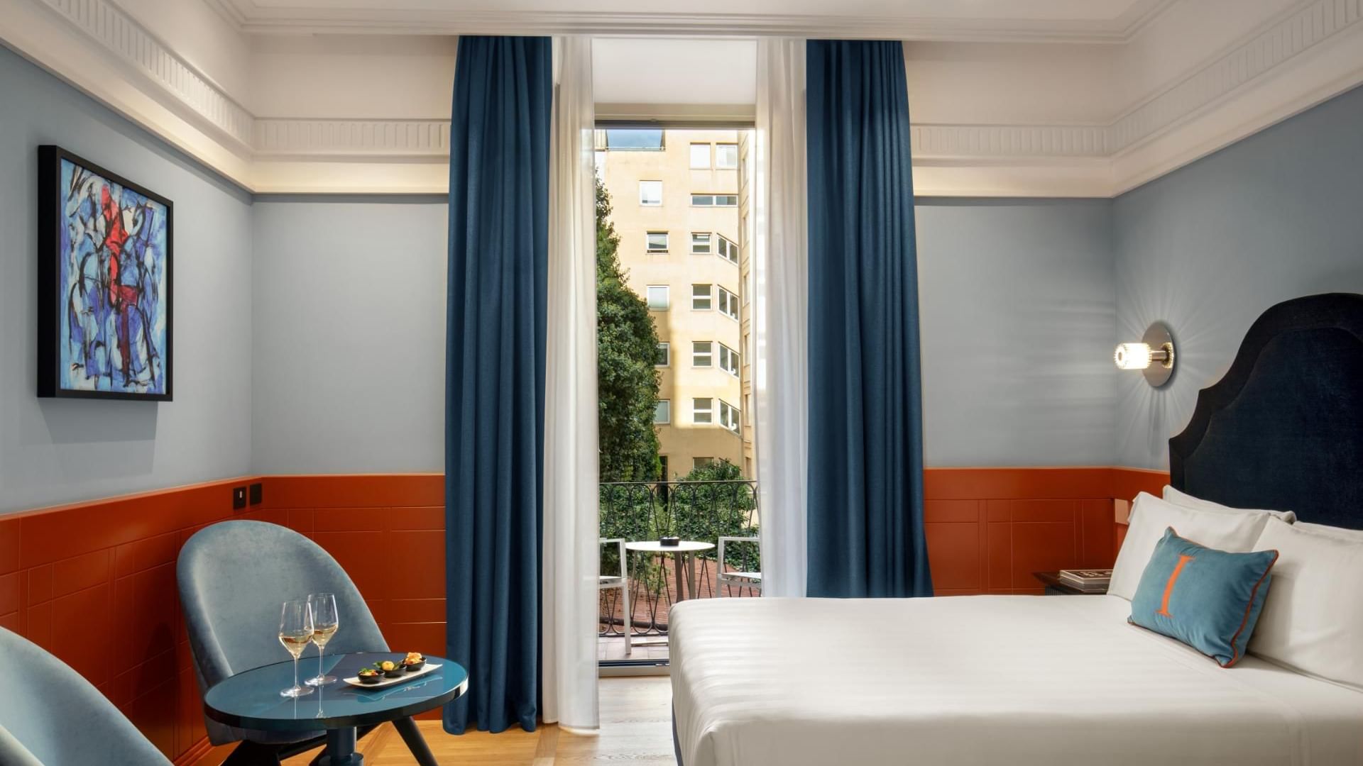 Bright open suite at The Independent with a double bed, a drink table, and a balcony view of buildings and greenery