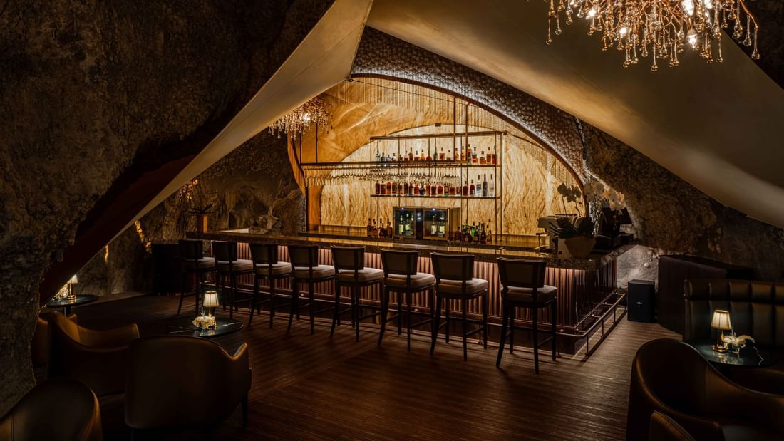 A dimly lit bar with a stone wall, wooden floor, and a large chandelier hanging from the ceiling.