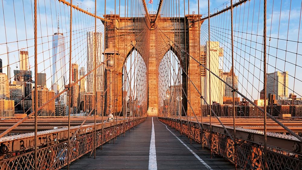 Long view of Brooklyn Bridge leading to New York City skyline at sunset.