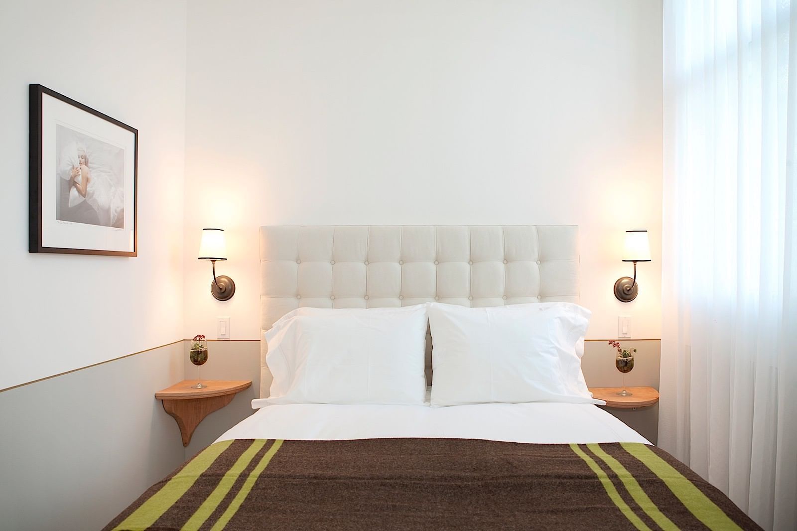 A neatly made bed with white pillows and a brown blanket at the foot, flanked by two wooden nightstands with lamps, and a framed picture on the wall.
