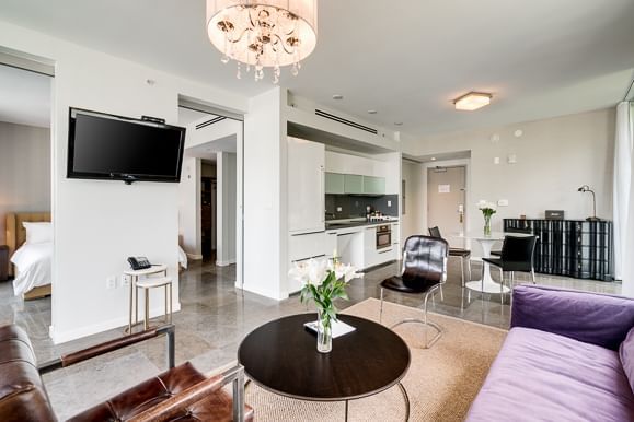 A wide view of a modern hotel suite with a kitchenette, seating area, bedroom, and TV.