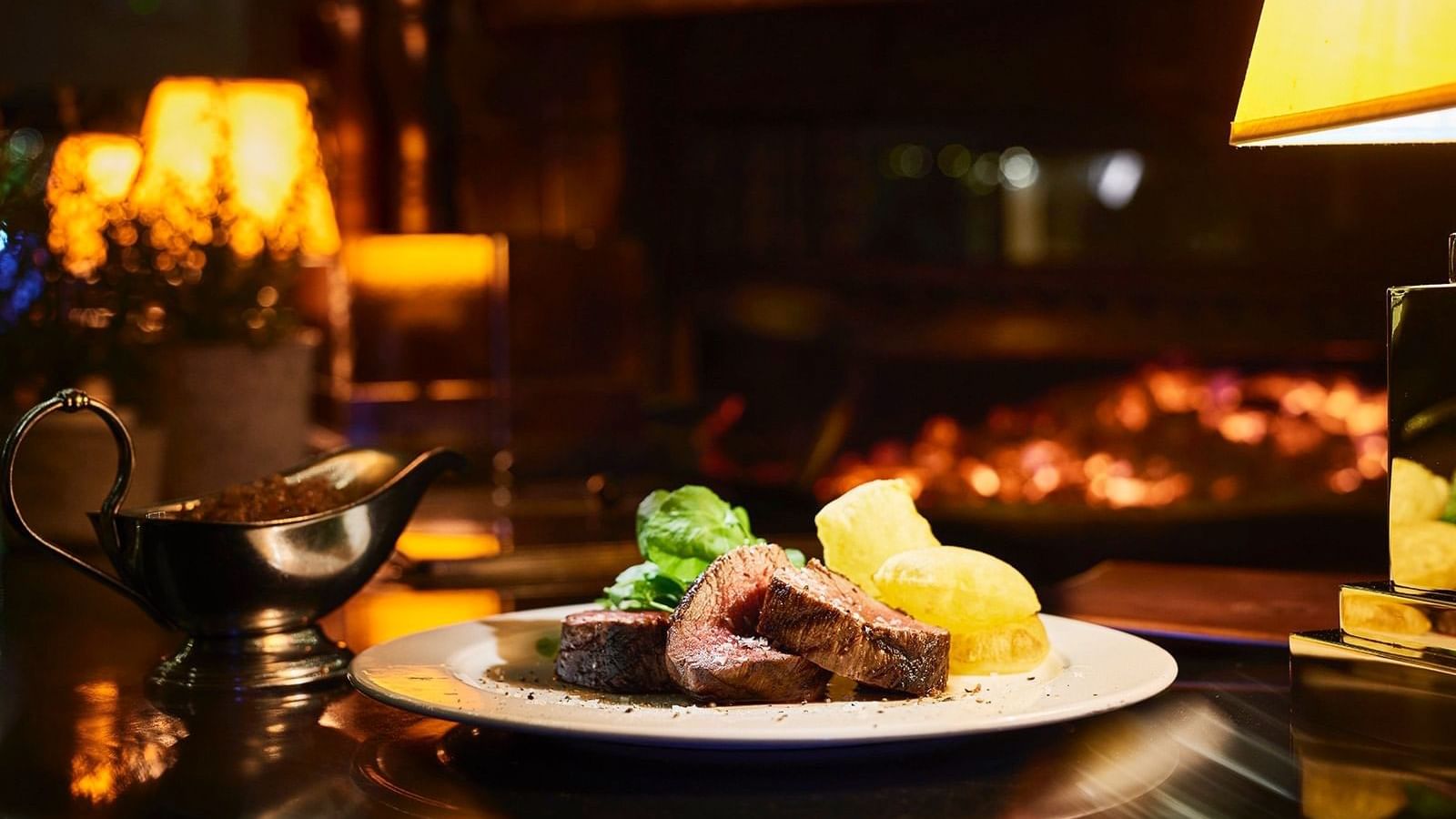 Plate with sliced roasted meat and potatoes near a gravy boat, with a fireplace in the background at the Marbella Club