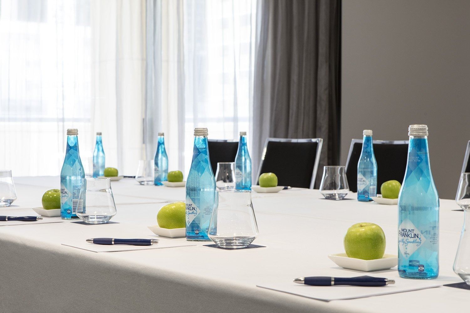 A bright meeting room table set with individual water bottles, green apples, and pens, ready at The Sebel Brisbane