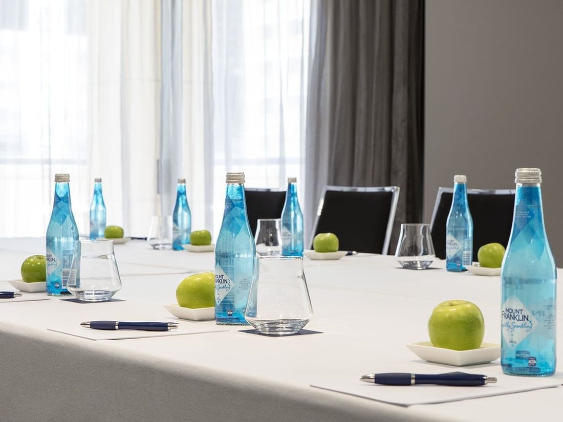 A bright meeting room table set with individual water bottles, green apples, and pens, ready at The Sebel Brisbane