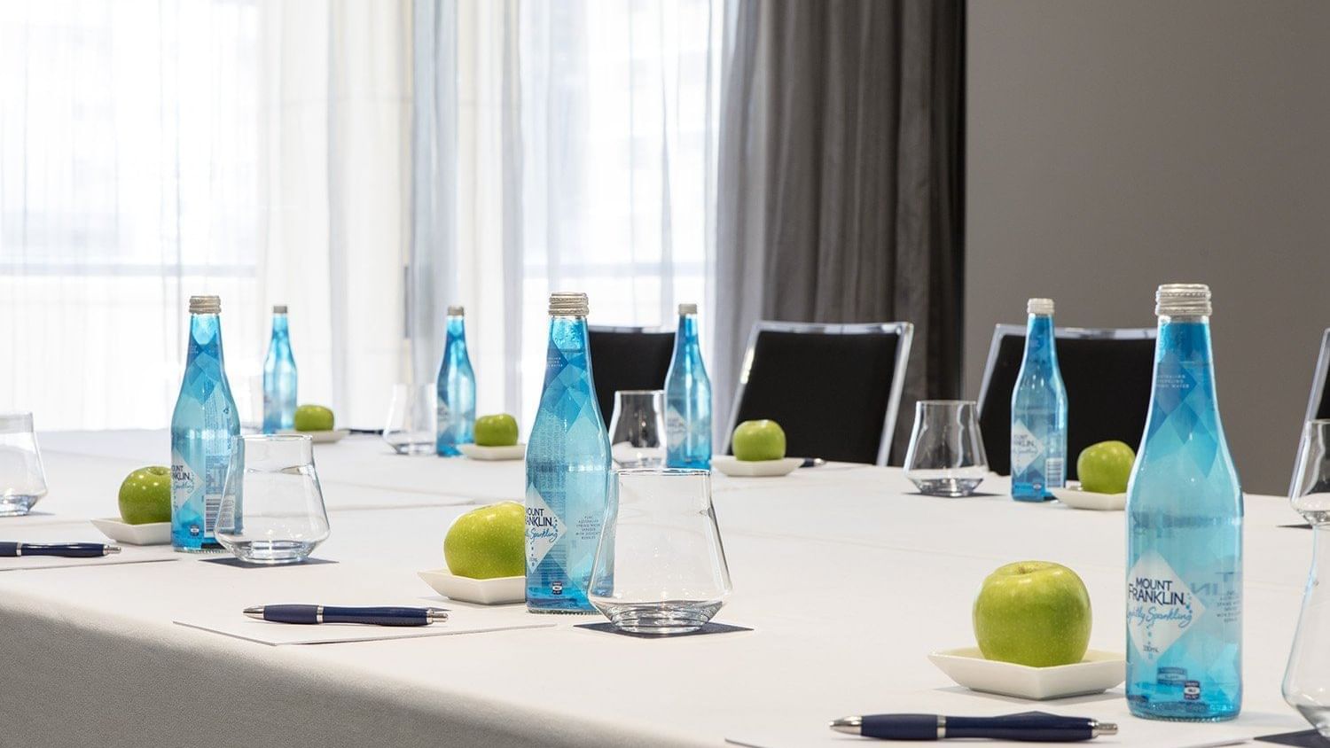 A bright meeting room table set with individual water bottles, green apples, and pens, ready at The Sebel Brisbane