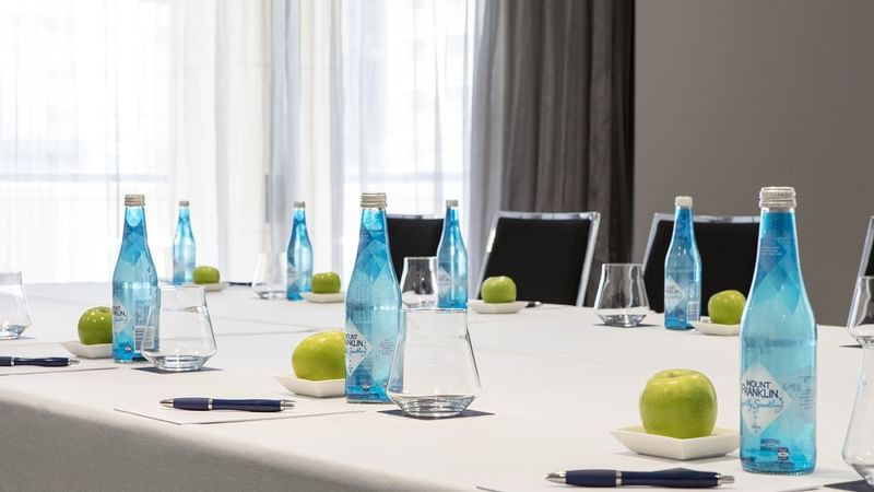 A bright meeting room table set with individual water bottles, green apples, and pens, ready at The Sebel Brisbane