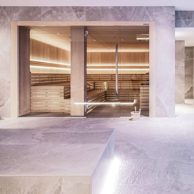 Moderne Sauna mit hellen Holzbänken und Glastüren, neben einem gefliesten Ruhebereich.