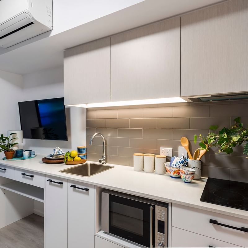 Modern studio kitchenette with white cabinetry, stainless steel sink, and decorative plants at UniLodge School Street Studios.