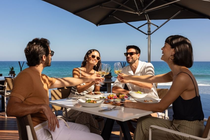 Four guests toasting with white wine at an oceanfront dining table under an umbrella at The Club at Solaz