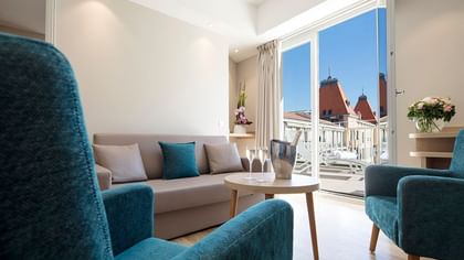 Living area with a sofa, two blue armchairs, and a balcony featuring city roof views from the Splendid Hotel & Spa Nice