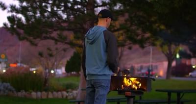 A man grills over an open flame at dusk, surrounded by green trees and warm evening light near Moab Valley Inn