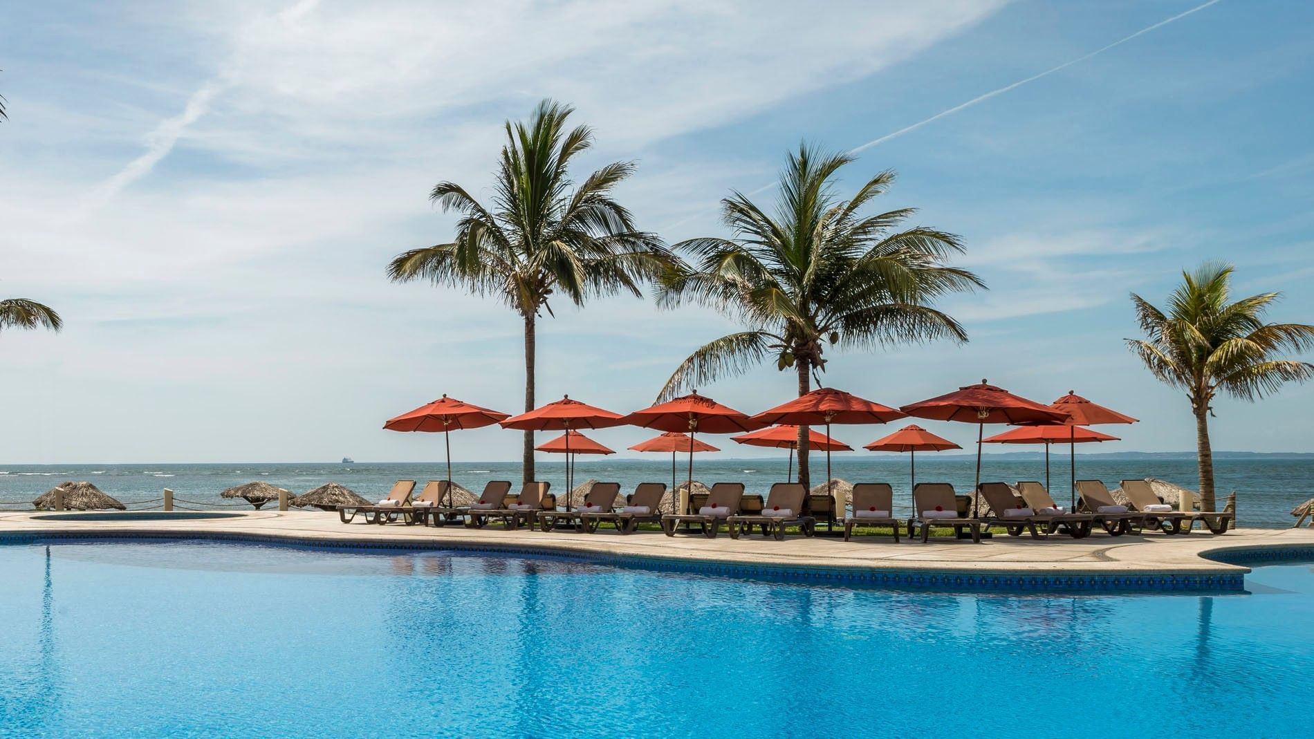 Pool Bar featuring blue swimming pool lined with red umbrellas and palm trees under a sunny sky at Camino Real Veracruz