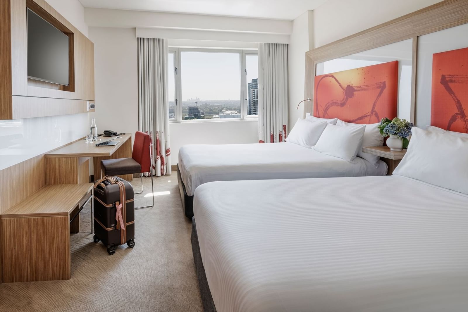 Room with warm decor featuring two kings beds, a desk, TV, and a window with a city view at Novotel Sydney Olympic Park