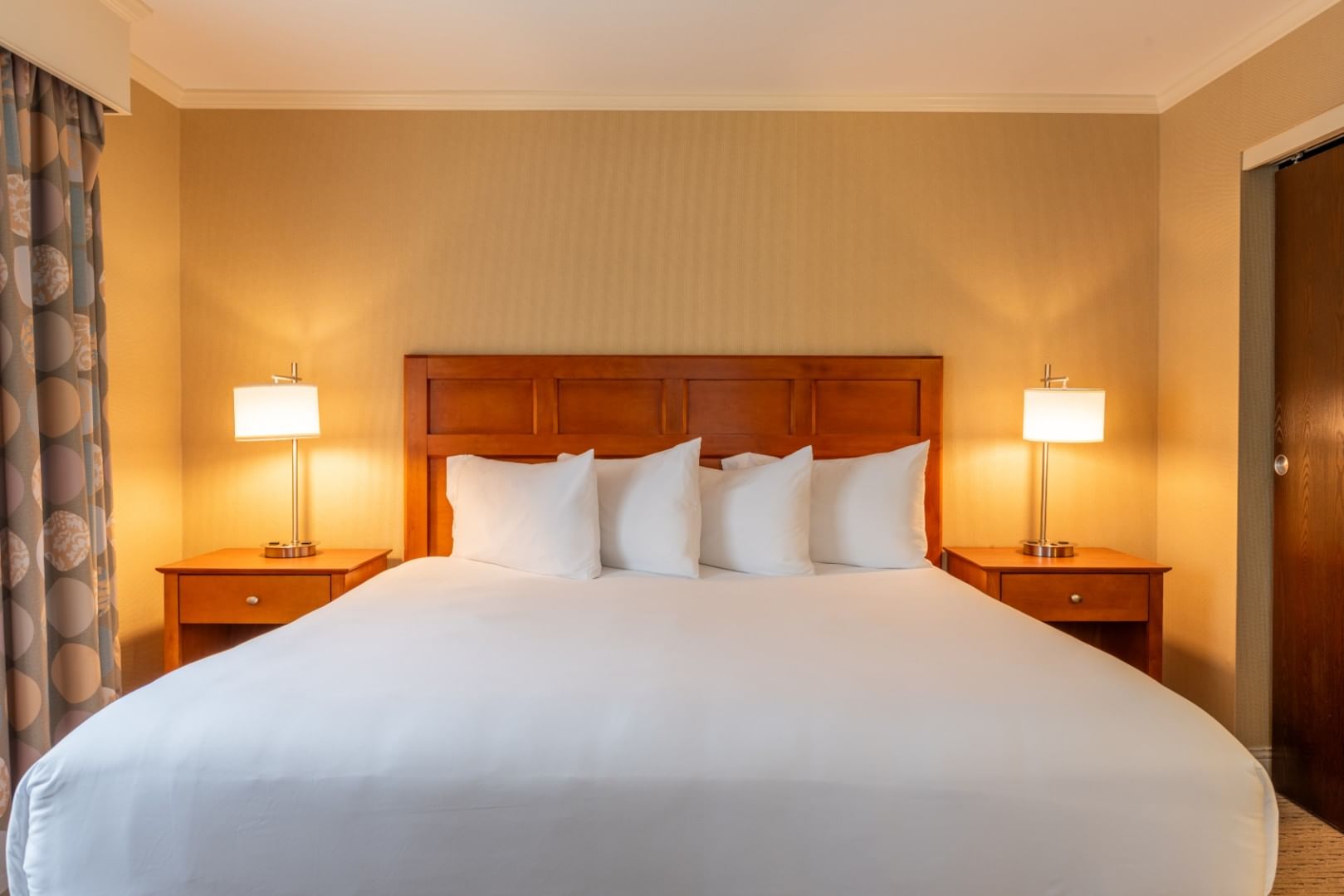 A wide view of a hotel room with a king-sized bed, wood headboard, two nightstands with lamps, and neutral-colored walls and bedding.