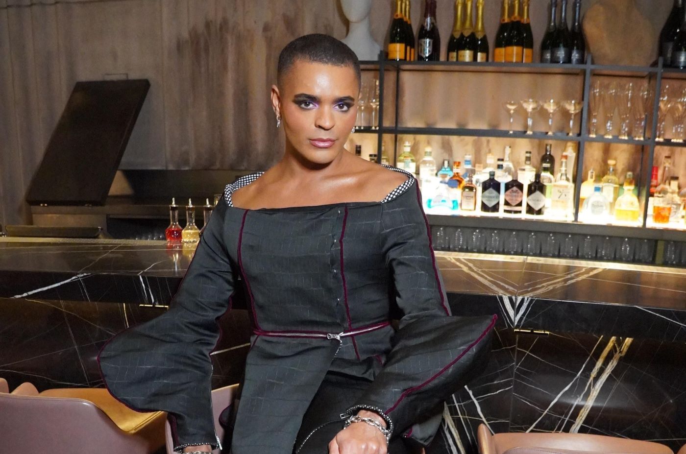 A person in a chic black top posing at a bar with various liquor bottles in the background at The Londoner Hotel