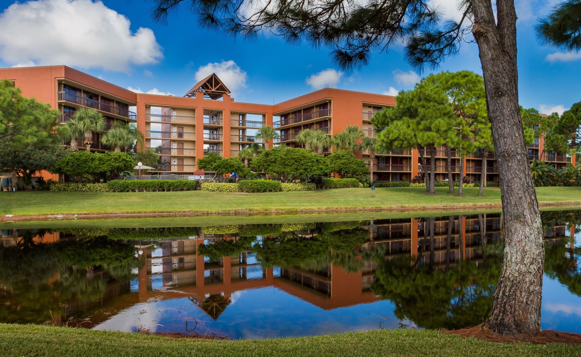 A beautiful burnt umber colored hotel surrounded by trees, green grass, and a reflective lake. Stay at a Rosen Inn to be close to the best immersive dining in Orlando.