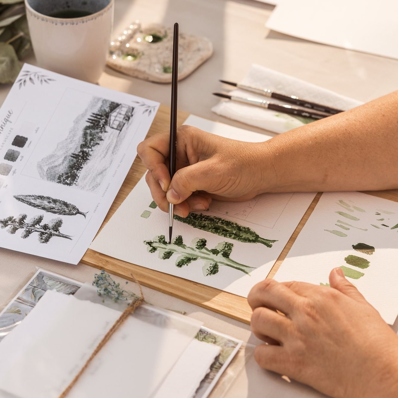 Taller de pintura botánica con acuarelas sobre papel, pinceles y plantas naturales sobre mesa de madera clara.