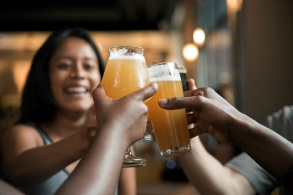 A group of hands raise their beer glasses in a toast with a woman smiling and out of focus in the background. 