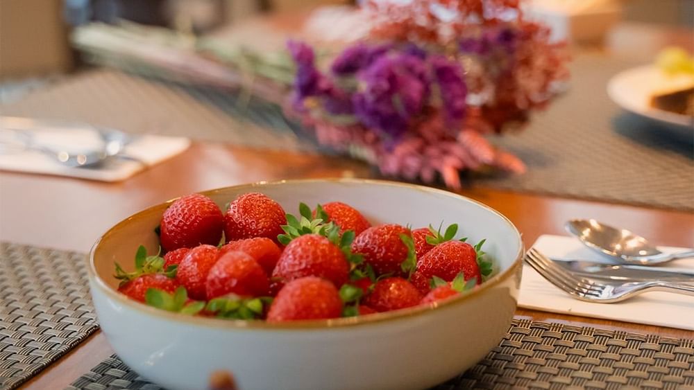 Un bol de fraises rouges et des couverts sur une table à manger chez Warwick Hotels and Resorts, avec des fleurs floues en arrière-plan