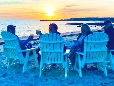 A group of guests sitting on outdoor chairs by the beach & enjoying beer during sunset near Chatham Tides Resort