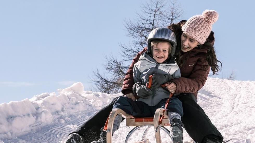 A child and an adult enjoy sledding down a snowy hill near Falkensteiner Hotels & Residences