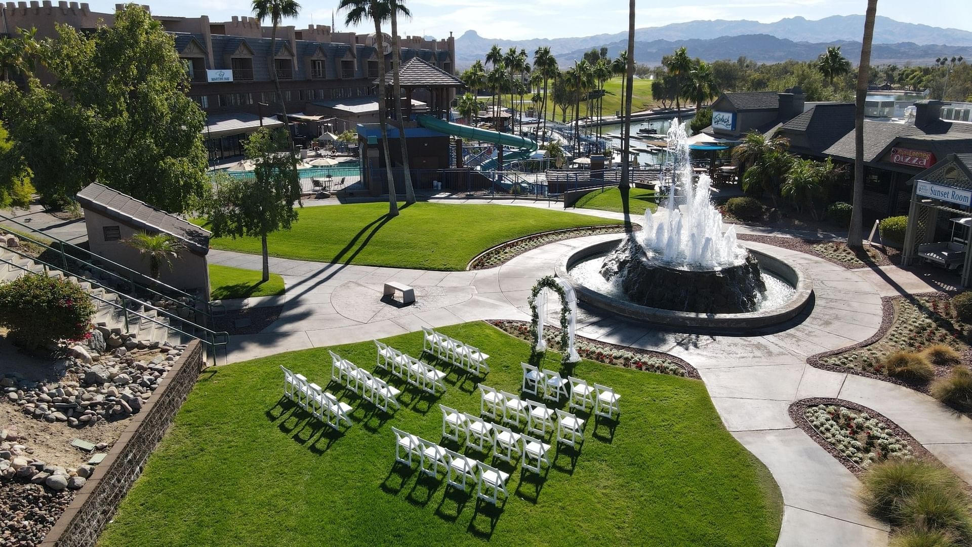 Outdoor wedding ceremony setup with white chairs on green lawn beside fountain and pool at London Bridge Resort