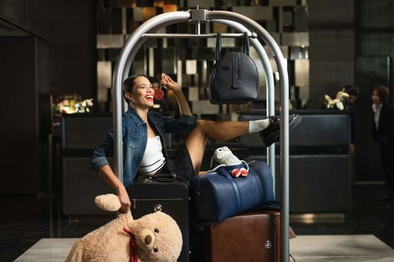 Woman sitting on luggage cart at a Paradox Hotel lobby, holding teddy bear and lollipop, with bags and a purse hanging above.