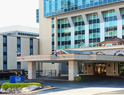 Porte-cochere entrance with flags and glass canopy facing driveway with blue property sign at Clayton Plaza Hotel