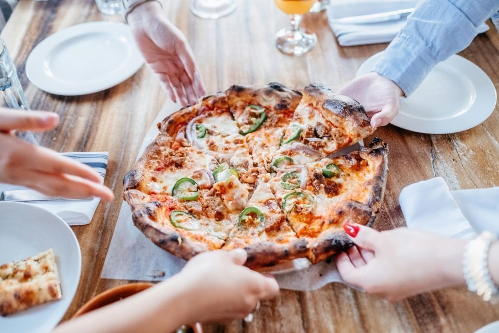 Five hands grabbing slices of pizza at the center of a wooden table surrounded by plates and glasses.