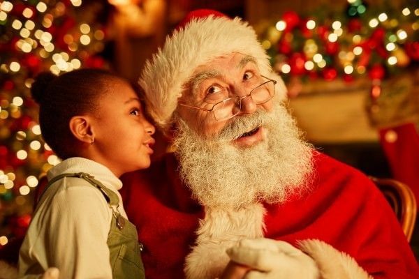 A little girl meeting Santa and whispering into his ear, surrounded by twinkling Christmas lights.