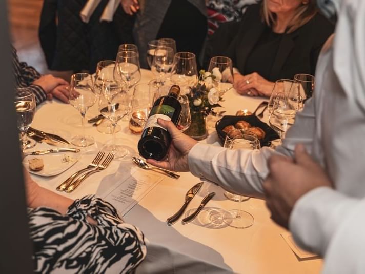 Hand holding wine bottle above a table with food, wine glasses, and utensils with people seated around.