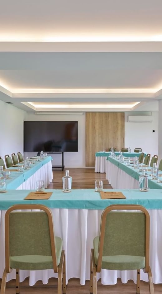 Conference room setup with green chairs around U-shaped teal tables facing a large display screen at Caloura Hotel Resort