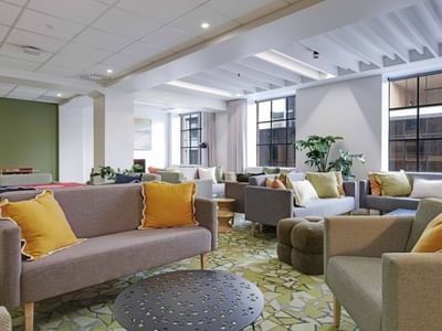 Modern communal lounge with gray sofas, yellow cushions, large windows, and potted plants at UniLodge Auckland Central.