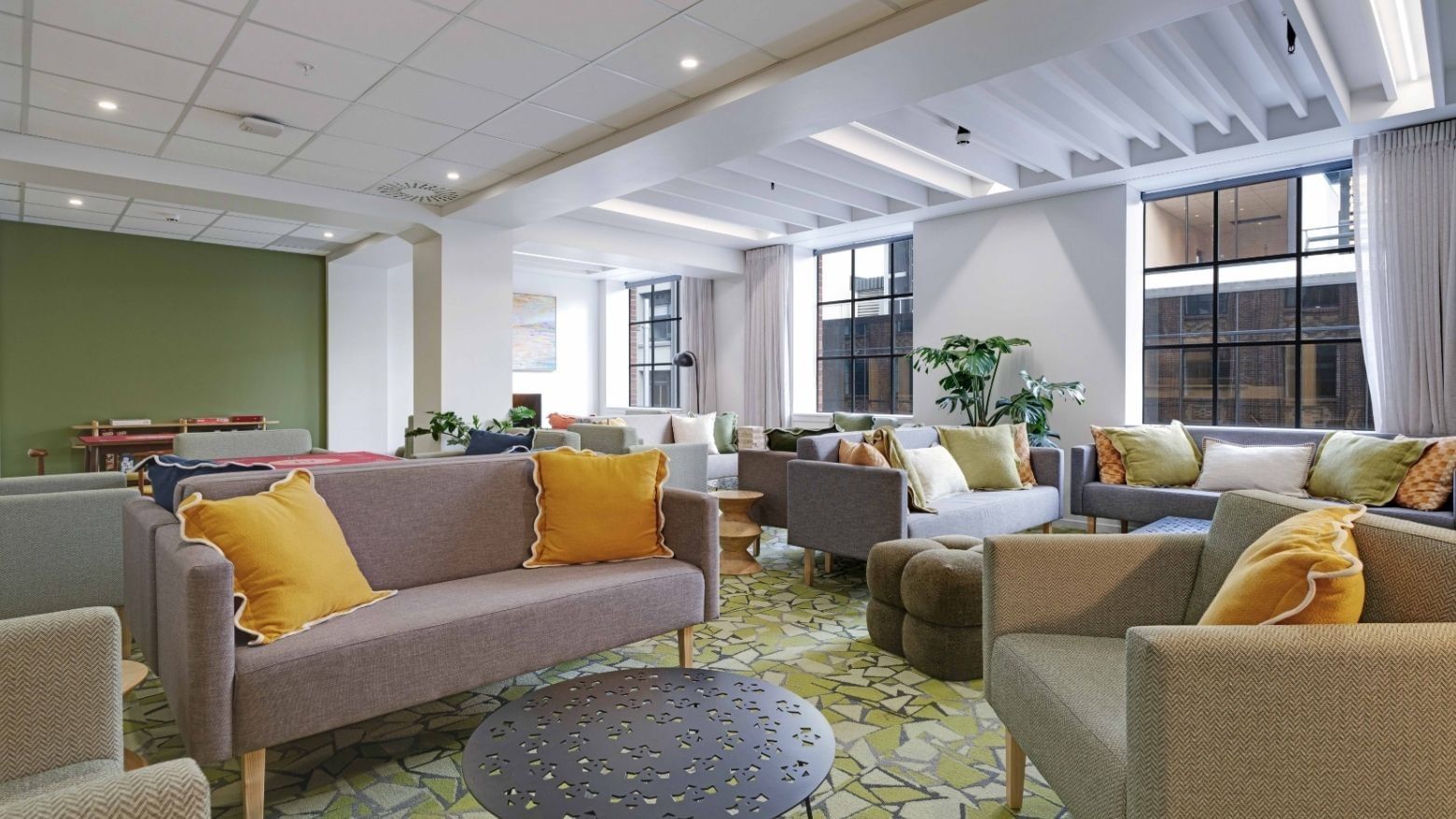 Modern communal lounge with gray sofas, yellow cushions, large windows, and potted plants at UniLodge Auckland Central.