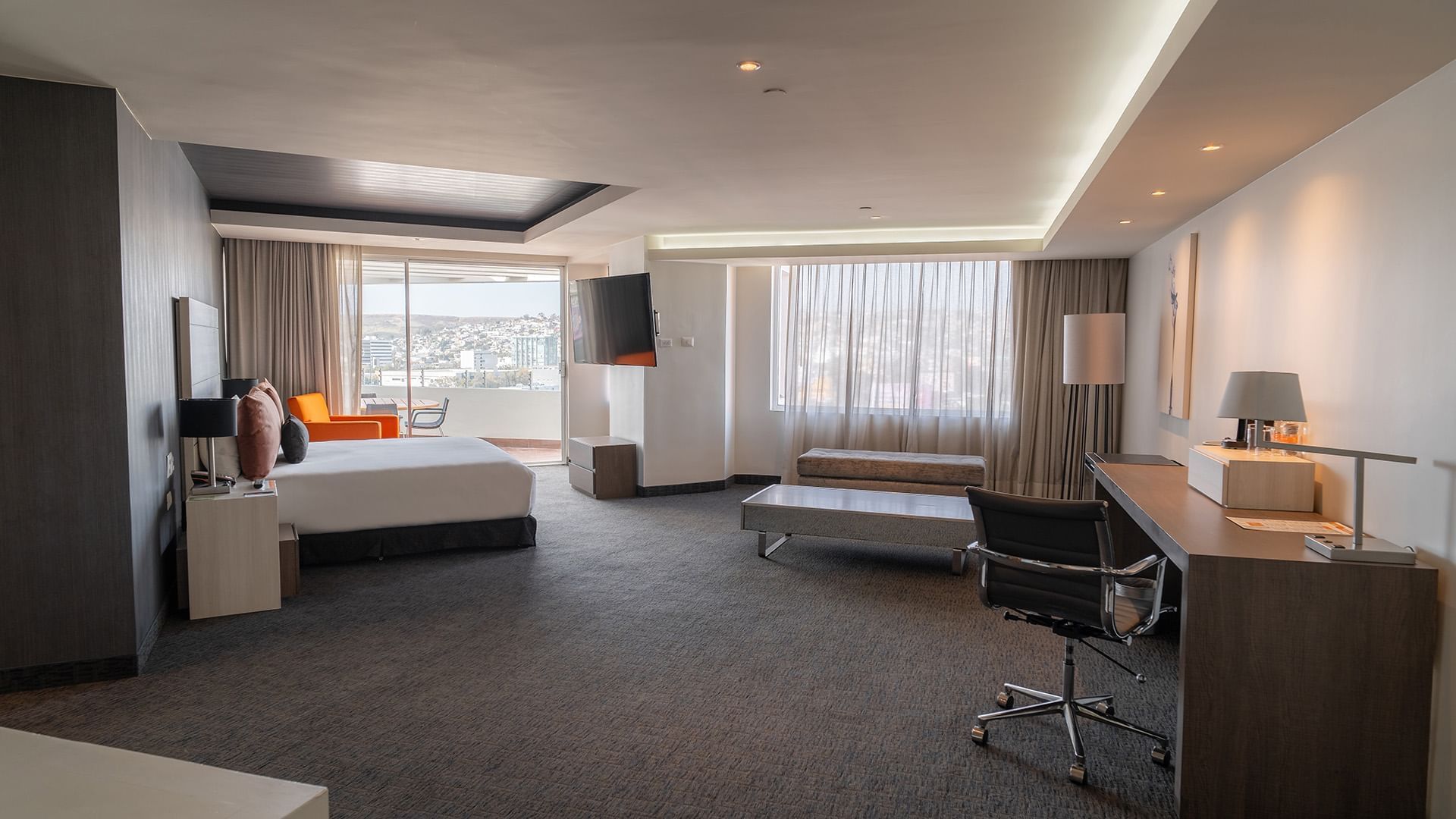 TV, cozy seating area, and work desk by the bed in the King Presidential Suite with carpeted floors at Real Inn Tijuana