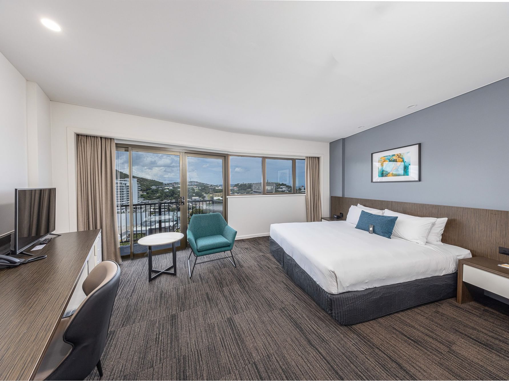 A spacious, modern hotel room featuring a large bed with white linens and a blue accent pillow against a blue feature wall. The room includes a long wooden desk with a TV, a teal armchair, and floor-to-ceiling glass doors opening onto a balcony with an ex