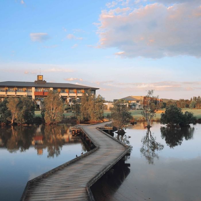 Mercure Kooindah Waters by a lake with a curving wooden footbridge and trees at sunset