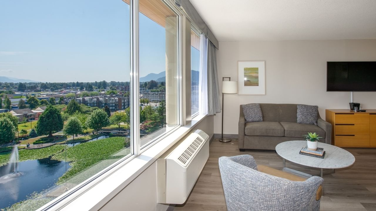 Hotel room living area with couch, chair, table, TV, and large window overlooking a pond.