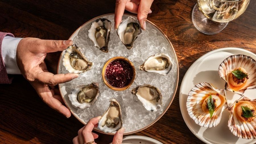 Hands reaching for fresh oysters on ice with mignonette sauce alongside seared scallops on a wooden table with white wine
