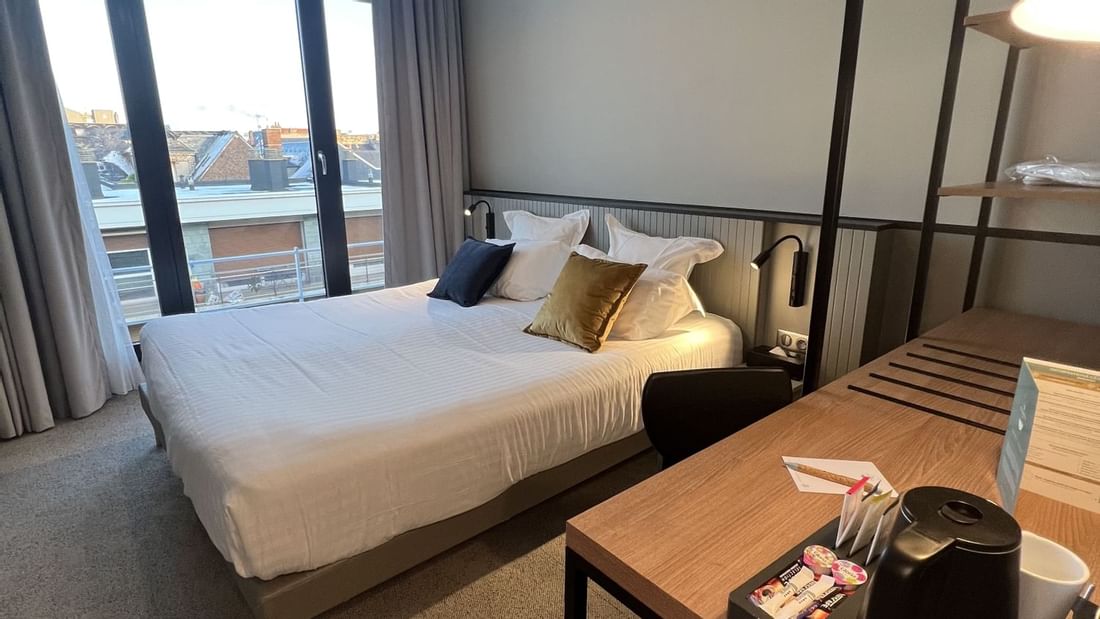Modern hotel room with bed, desk, chair, large window, and hanging lamps, overlooking cityscape.