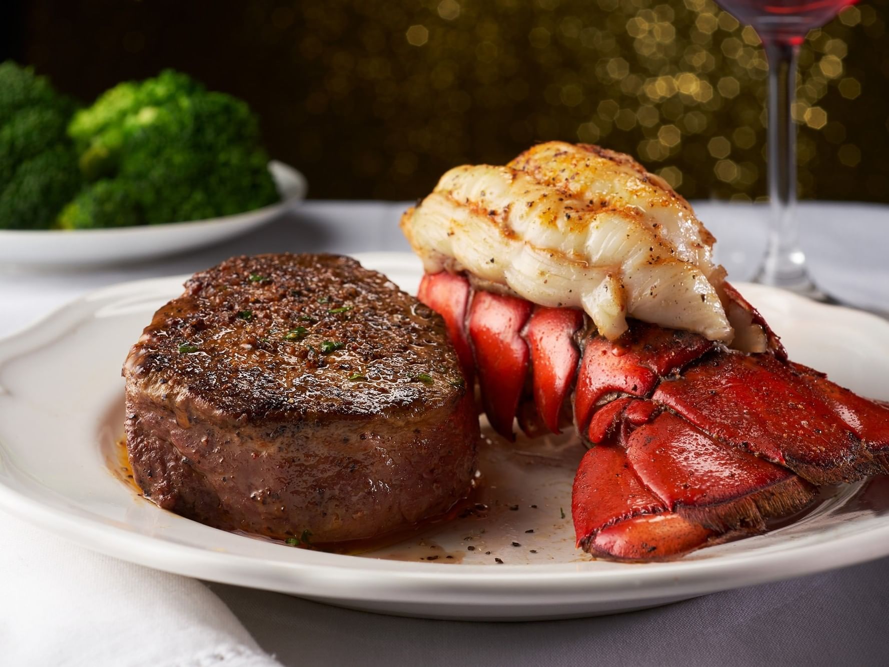 A plate of steak and lobster with a side of broccoli and a glass of wine