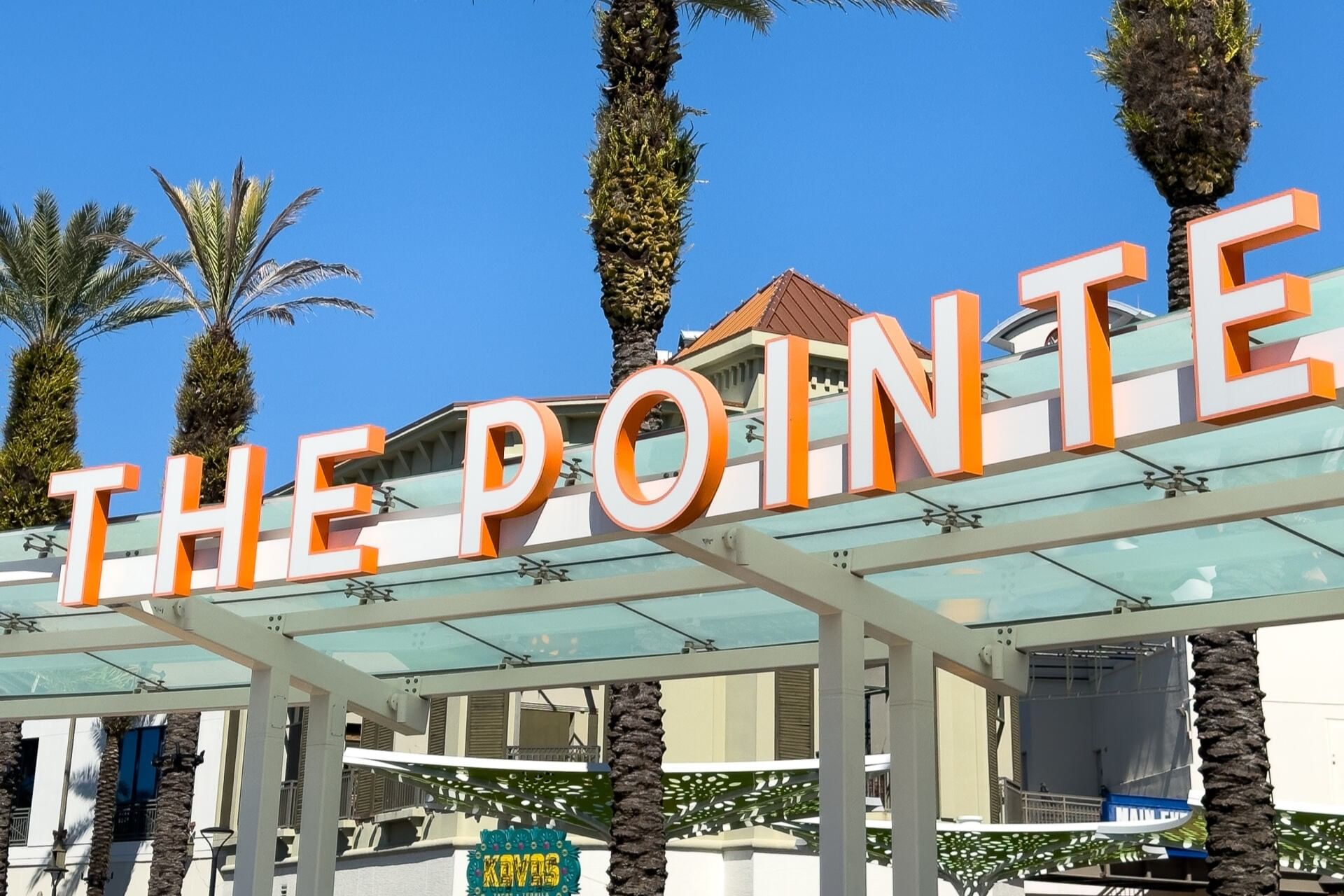 White and orange letters spelling The Pointe stretch across an awning surrounded by blue sky and palm trees. 