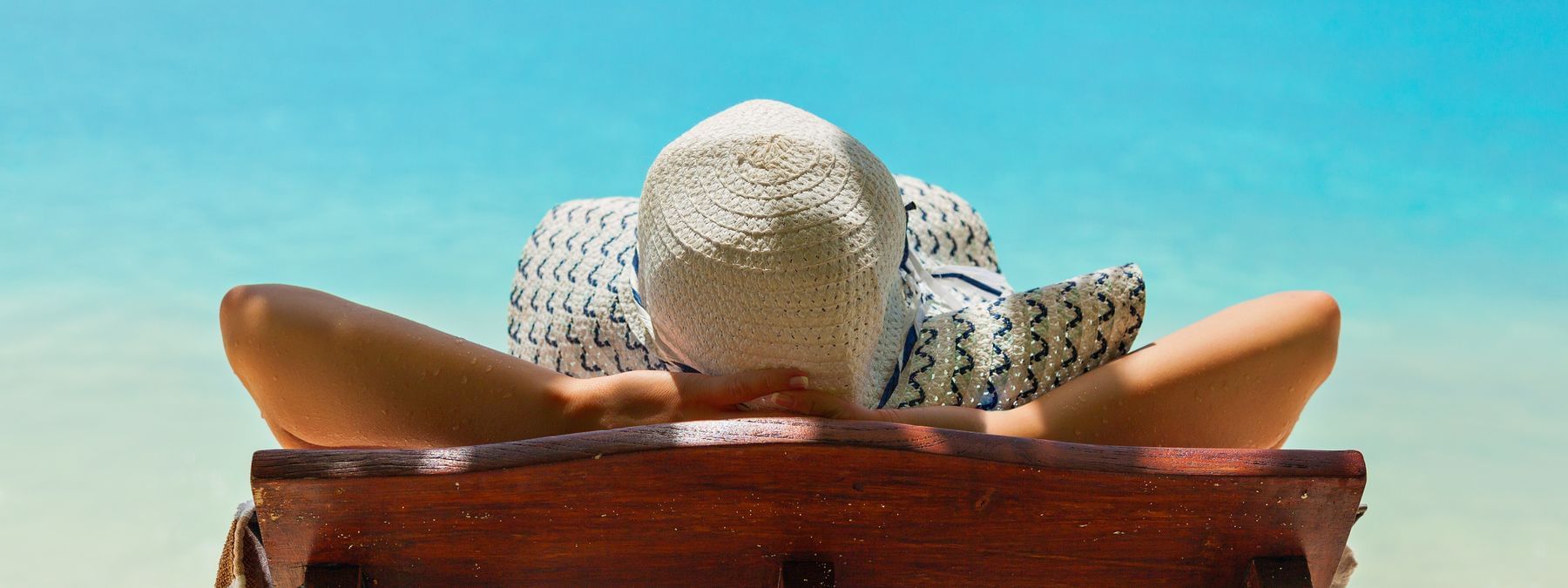 A woman in a hat relaxes on a wooden lounge chair at the beach with blue water and white sand in the background.