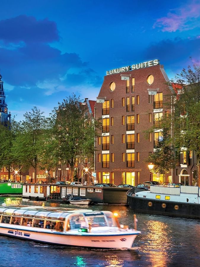 Luxury Suites Amsterdam exterior by the canal with tour boat & historic towers under sunset sky near Hotel Barsey by Warwick