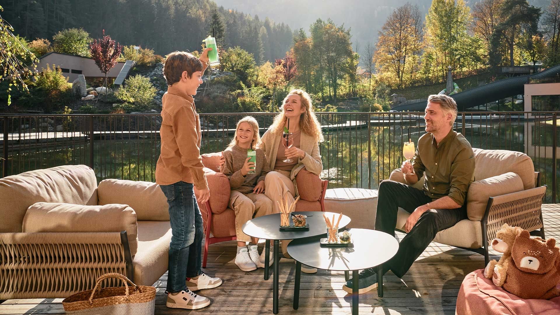 Family of four enjoying drinks and snacks on a balcony overlooking a park with trees and a water feature.