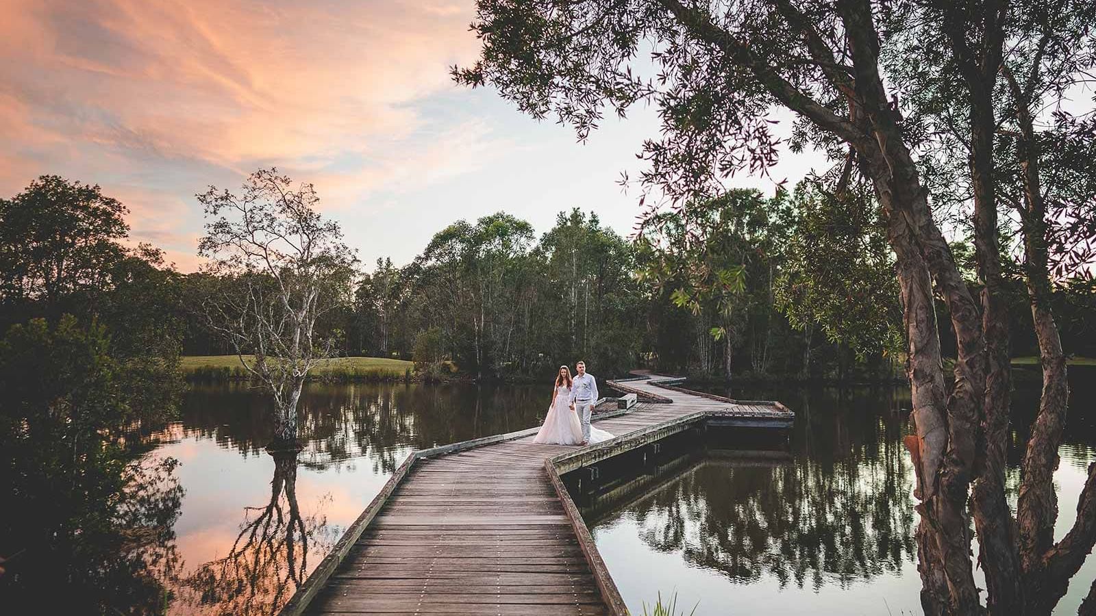 A bride and groom walking on a wooden pier by a lake near Mercure Kooindah Waters