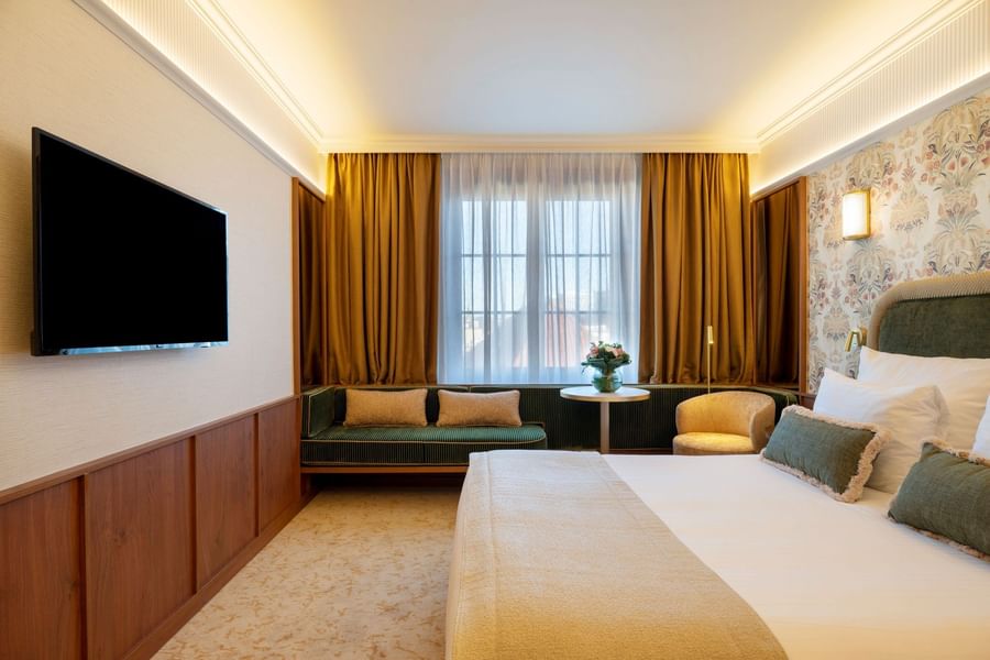 Elegant hotel room with white bed, green and gold accents, window seating, and wall-mounted TV at Warwick Brussels Grand-Place