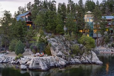 A lake with a group of people by a fire surrounded by rocks and trees with houses in the background.