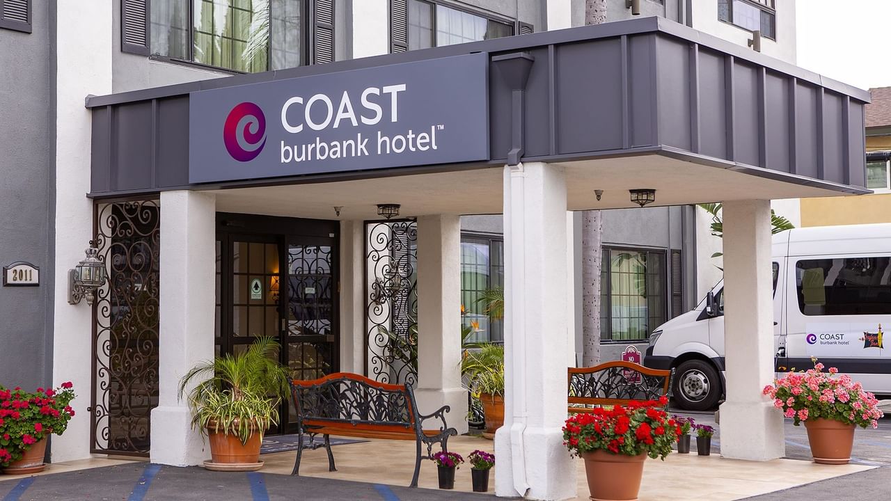 Entrance of Coast Burbank Hotel with benches, potted plants, and a van parked nearby.